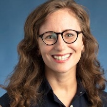 Woman with brown wavy hair and glasses smiling blue background