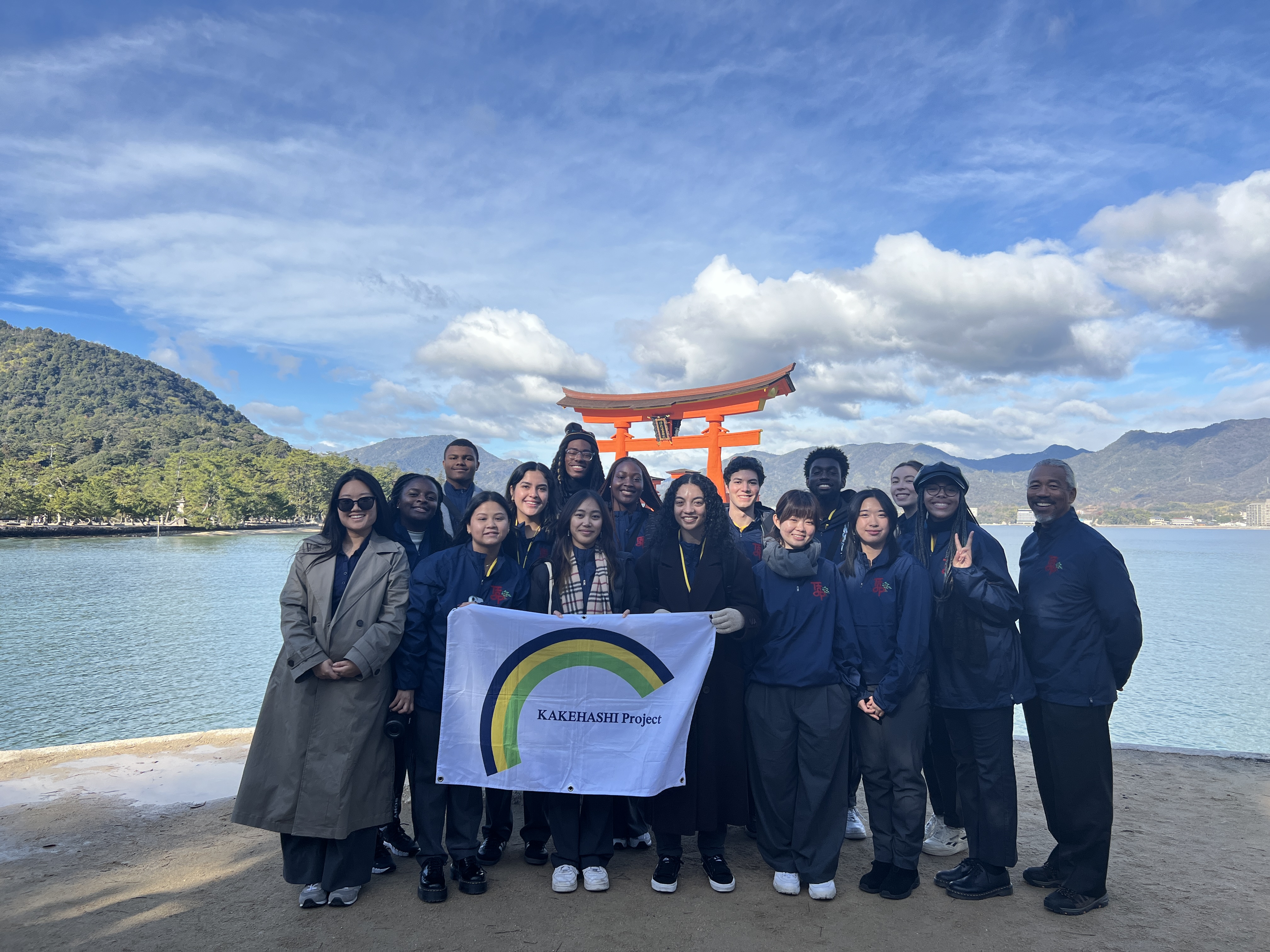 14 LMU students along with Dr. Curtiss Takada Rooks and Min-Jung Kim in front of the Itsukushima Shrine in Japan while holding a white banner that reads KAKEHASHI PROJECT