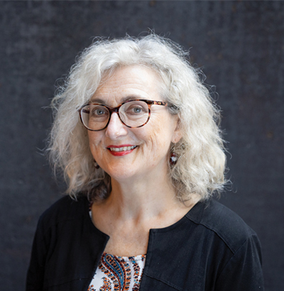 Grey haired woman wearing glasses, black sweater posing in front of a dark background